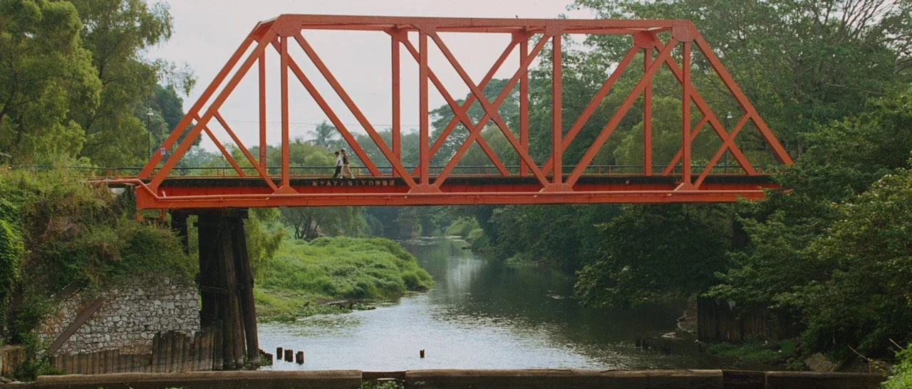 Sin Nombre (2009) movie still: wide — A bright orange steel truss bridge crosses over a slow flowing river. Two people are walking slowly along the…