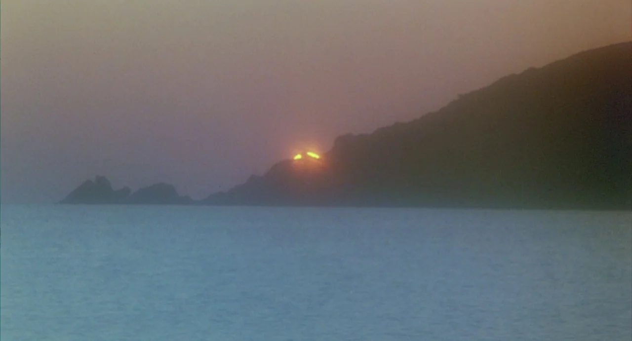Conquest (1983) movie still: establishing — A coastline featuring a dark mountain silhouette against a sunset sky. The sun shines brightly over the…