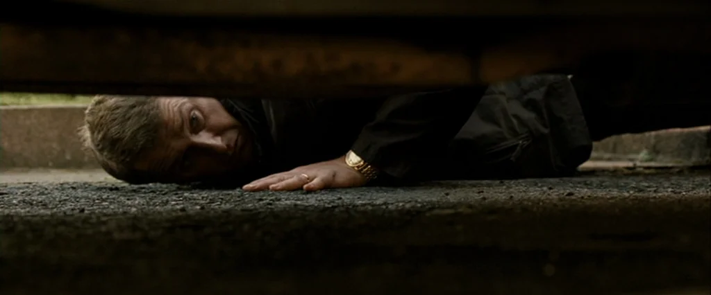 Hunger (2008) movie still: closeup — A man lies flat on a dark asphalt surface beneath the undercarriage of a vehicle. The low angle perspective…