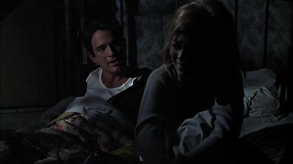 Bonnie And Clyde (1967) movie still: medium — A dimly lit room with floral wallpaper and a metal-framed bed at night. A young man and woman sit in…
