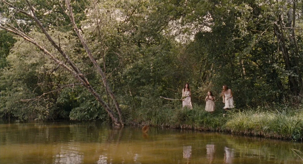 House of Tolerance (2011) movie still: wide — Three women in white dresses stand by a riverbank surrounded by dense foliage. A large bare tree trunk…