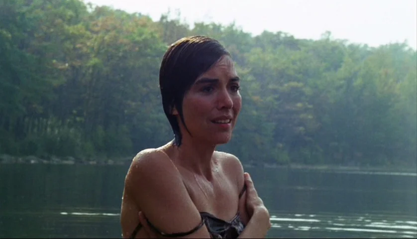 Let’s Scare Jessica To Death (1971) movie still: medium — A woman stands in a lake surrounded by a dense forest under overcast skies. She keeps her…