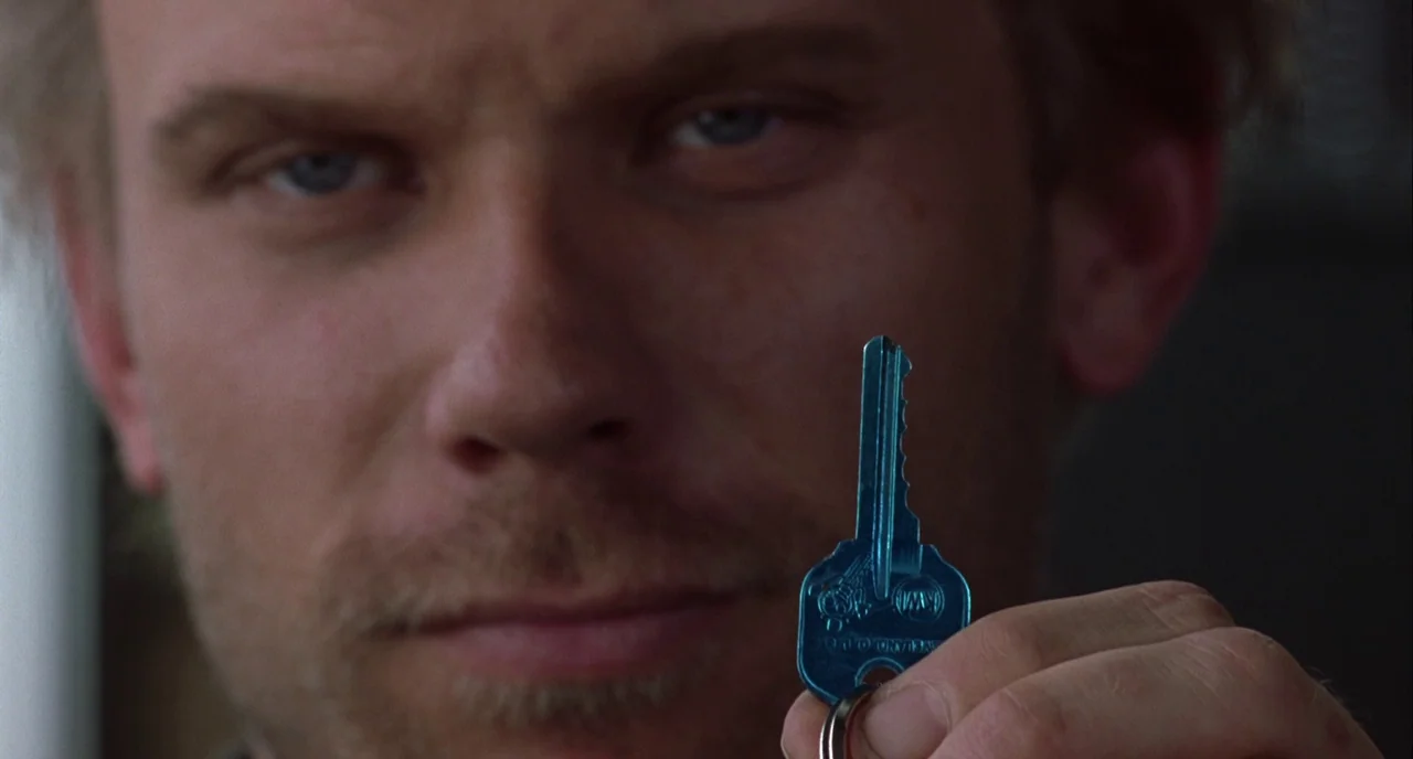 Mulholland Drive (2001) movie still: closeup — A man holds a small blue key in the foreground of a blurred room. The key is in sharp focus while the…