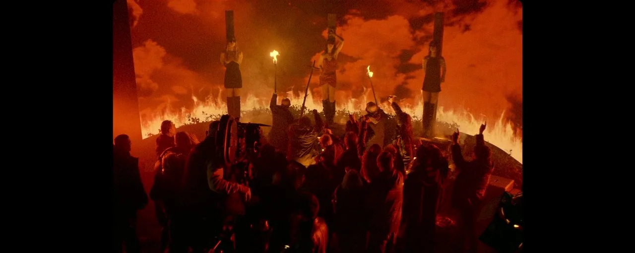 Lux Æterna (2019) movie still: wide — A film crew surrounds three women tied to posts in front of a ring of fire. The crew holds equipment and…