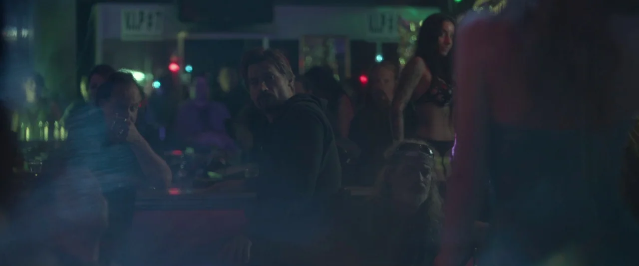 Small Crimes (2017) movie still: medium — A dimly lit club interior features a dark wooden counter with patrons and signage. A man in a dark hoodie…