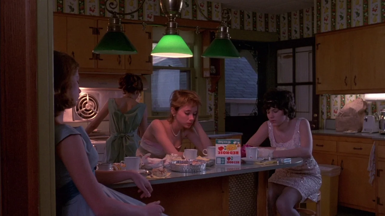 Baby It’s You (1983) movie still: medium — A 1950s kitchen features floral wallpaper, wooden cabinets, and green pendant lighting fixtures. Four…
