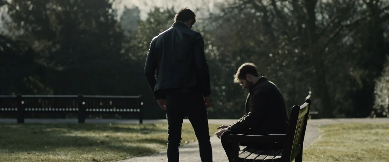 The Thorn On His Side (2014) movie still: fullbody — A public park path features a wooden bench under scattered trees during the day. Two men stand…