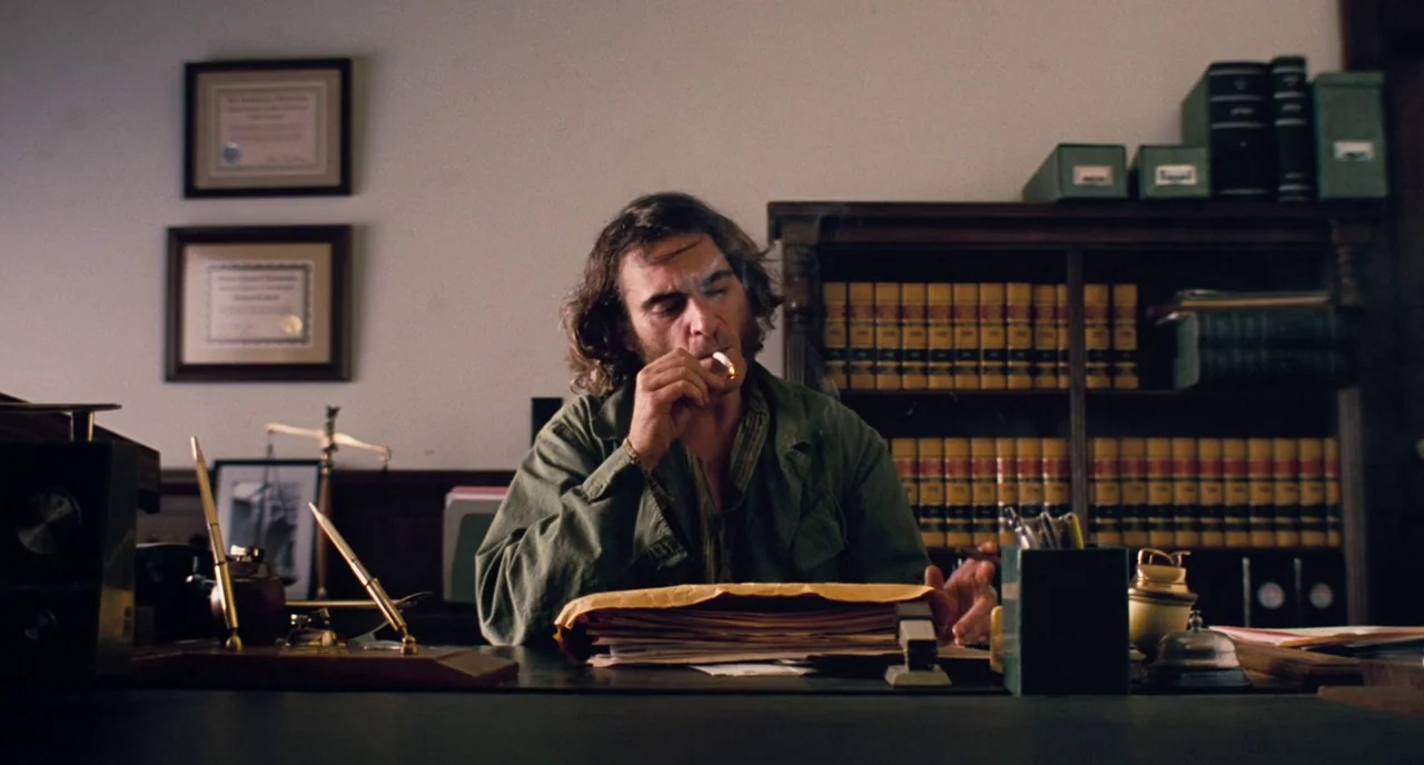 Inherent Vice (2014) movie still: medium — A cluttered 1970s workplace features dark wooden furniture, stacks of law books, and framed documents. A…