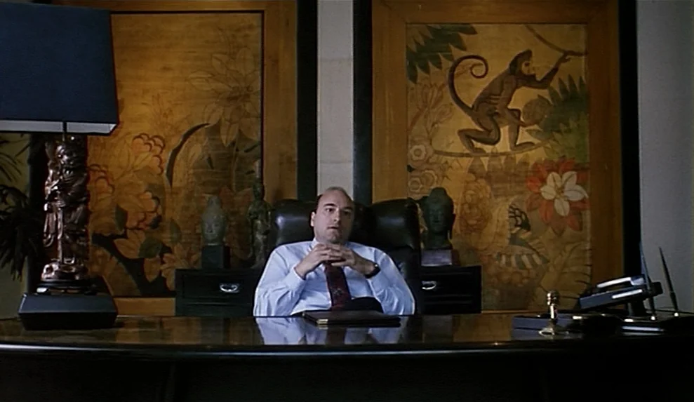 Safe movie still: medium — An office features two large framed paintings of jungle monkeys mounted on the wall. A man sits behind a dark wooden desk…