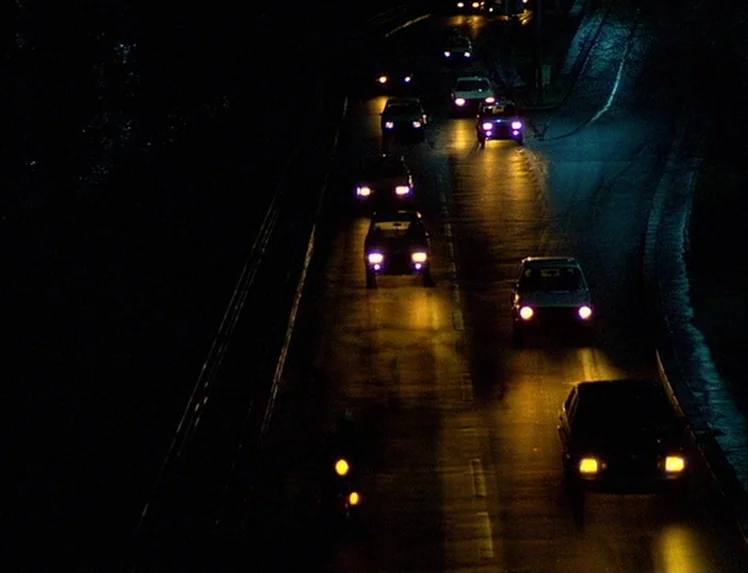 First Name: Carmen (1983) movie still: wide — A multi-lane asphalt road at night viewed from an elevated perspective. Several cars are moving along…