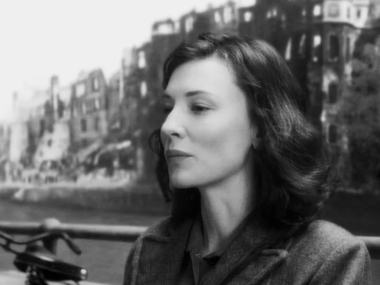 The Good German movie still: medium — A woman stands by a metal railing near a blurred waterfront cityscape backdrop. She stares off to the side…