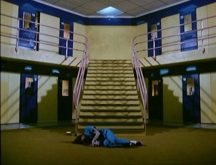 Ghosts… Of The Civil Dead (1988) movie still: wide — A symmetrical hallway features yellow walls and blue doors with numbered windows. A person in…