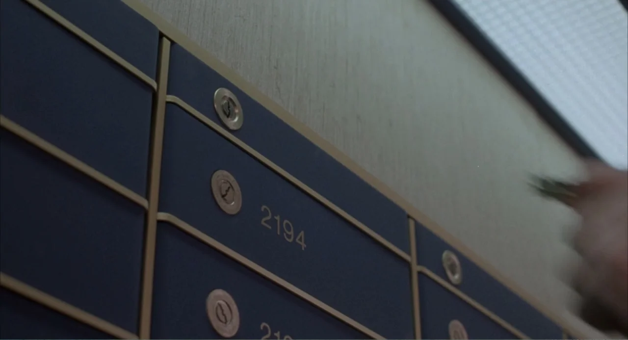 F/X (1986) movie still: closeup — A wall of numbered blue metal mailboxes with brass accents lines a corridor wall. A blurred hand moves across the…