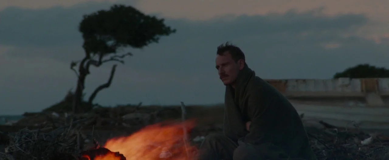 The Light Between Oceans (2016) movie still: medium — A coastal shoreline at dusk with a single wind-swept tree near the dark water. A man sits…