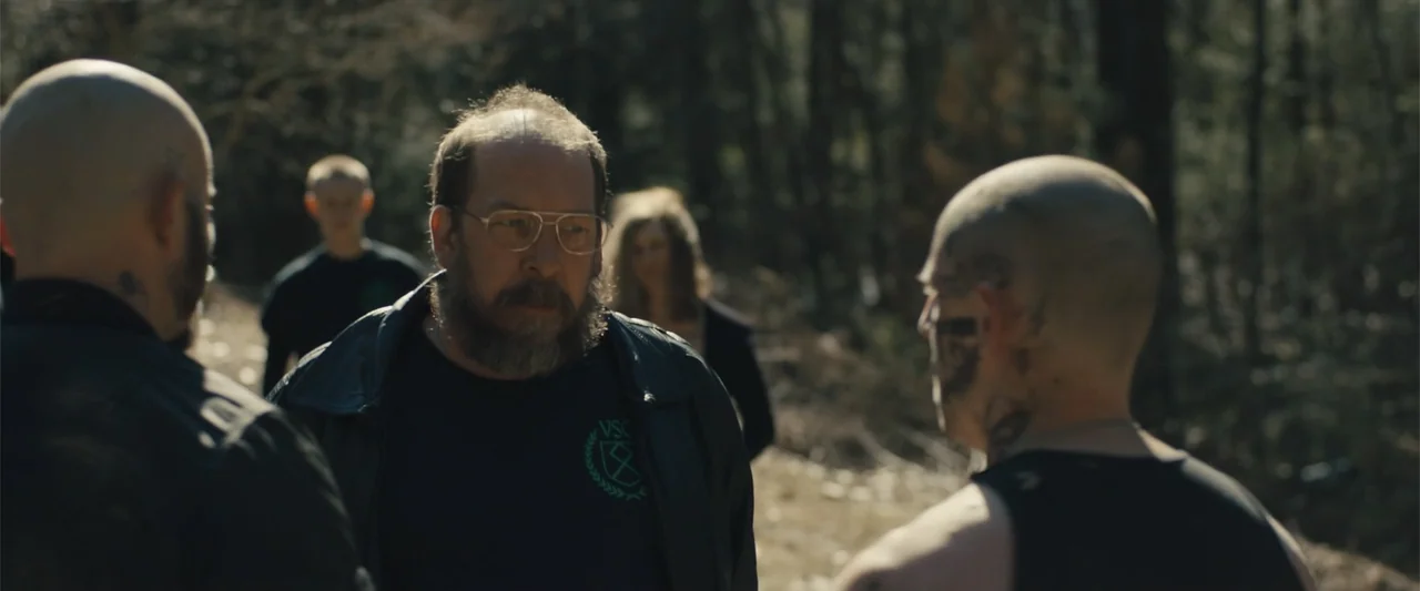 Skin (2009) movie still: medium — A group of people stands on a dirt path surrounded by dense forest trees. A man with glasses and a beard stands…
