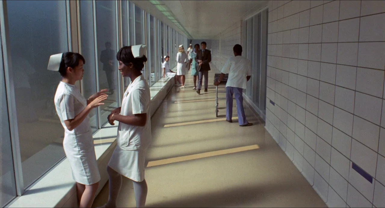 Medium Cool (1969) movie still: wide — A long hospital hallway features a glass wall with sunlight projecting shadows on the floor. Nurses in…