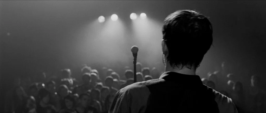 Control movie still: medium — A dark concert venue with a large audience visible in the out-of-focus background. A performer stands at a microphone…
