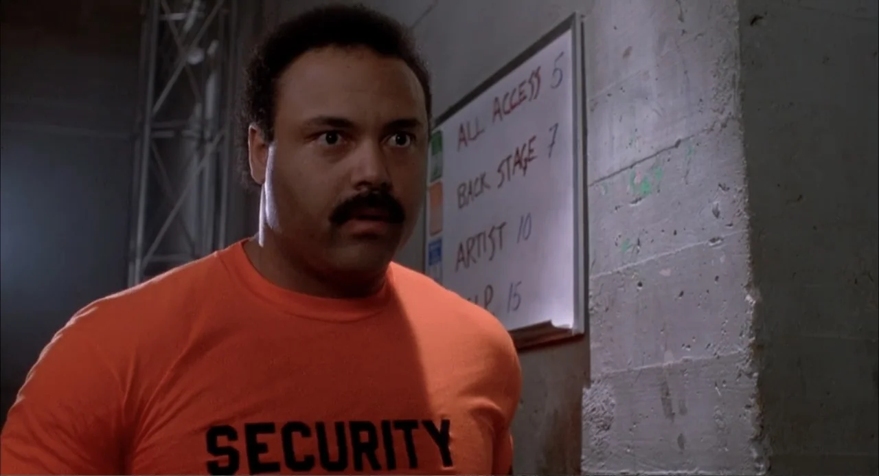 Teen Witch (1989) movie still: medium — A man in a security uniform stands in a dimly lit concrete corridor. He stares forward while standing next…
