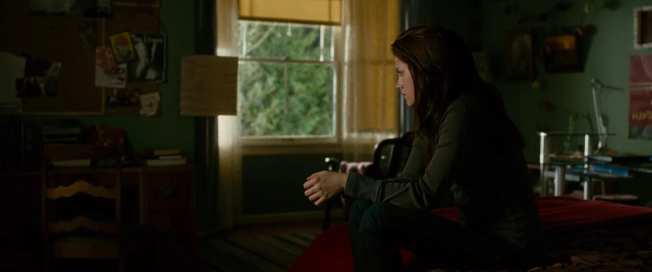 The Twilight Saga: New Moon (2009) movie still: medium — A dark bedroom has green walls covered in posters and a red bedspread. A young woman sits…