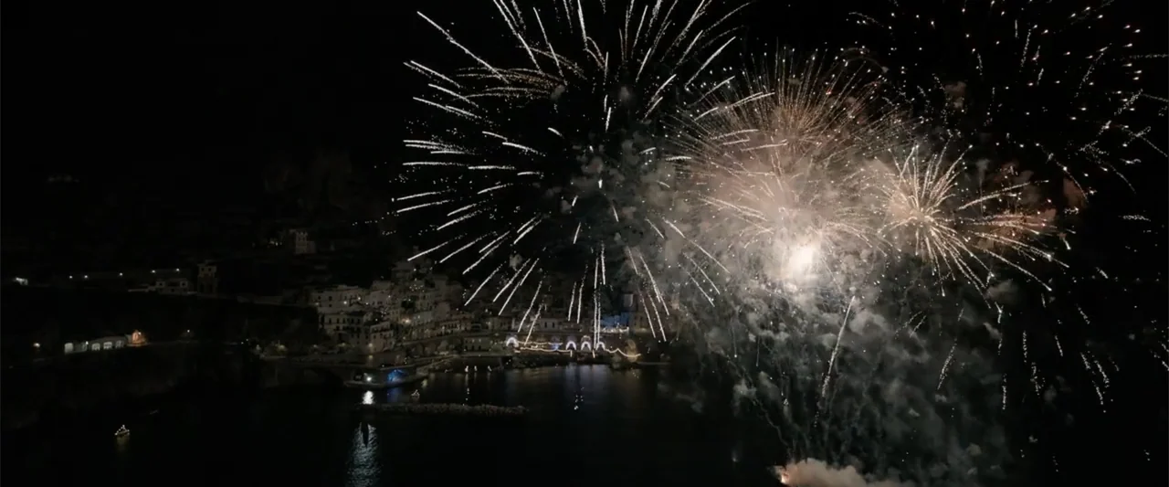 The Equalizer 3 (2023) movie still: establishing — Bright white fireworks explode above a dark coastal town situated along a calm harbor. The harbor…