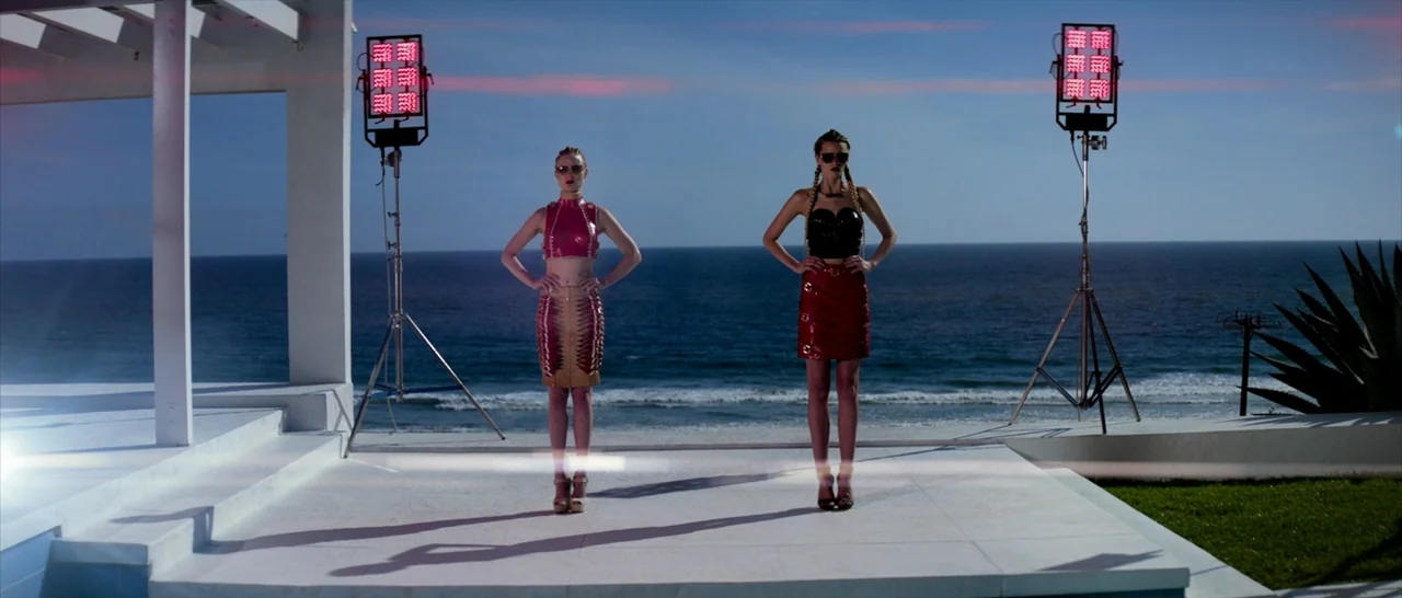 The Neon Demon (2016) movie still: fullbody — A white paved patio overlooks the ocean during a bright day. Two women stand side-by-side in front of…