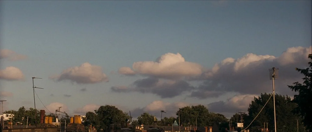 Happy-Go-Lucky (2008) movie still: establishing — A row of suburban chimneys stands beneath a wide expanse of partly cloudy sky. The horizon is…