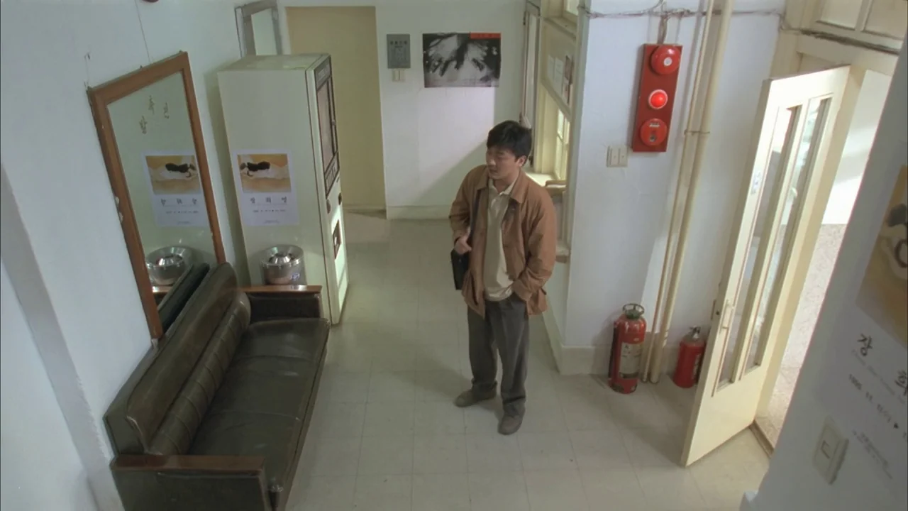 The Day a Pig Fell Into the Well (1996) movie still: fullbody — A sparse, brightly lit hallway with white walls and light tiled flooring. A lone man…
