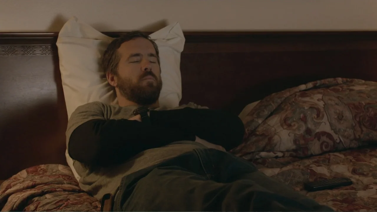 The Captive (2014) movie still: medium — A residential bedroom with a wooden headboard and patterned bedding. A bearded man lies on the bed with his…