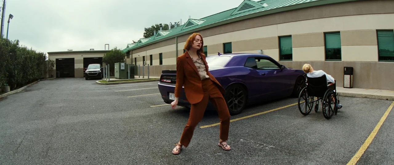Kinds of Kindness (2024) movie still: fullbody — A paved asphalt parking lot sits before a low, light-colored institutional building with green…