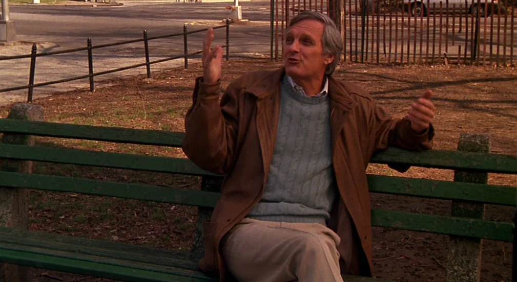 Crimes and Misdemeanors (1989) movie still: medium — A man sits on a weathered green wooden bench in a park setting. He gestures with his hands…