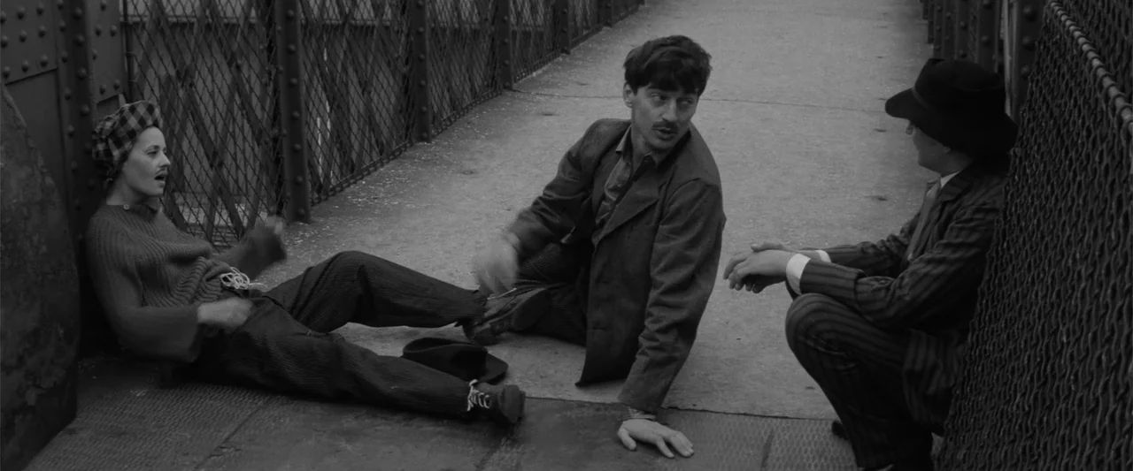Jules and Jim (1962) movie still: fullbody — Three people sit on the concrete path of a bridge enclosed by chain-link fencing. They are engaged in…