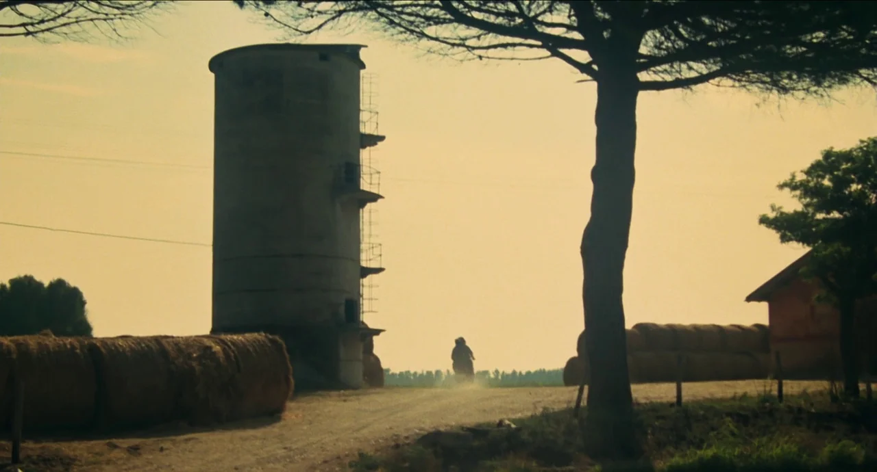 City of Women (1980) movie still: wide — A rural farmland scene features a tall cylindrical grain silo beside hay bales. A lone motorcyclist travels…