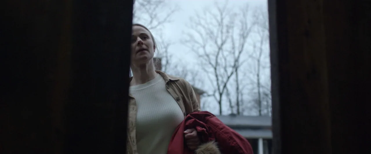 Motherly (2022) movie still: medium — A woman stands within a dark doorway looking out at a bare-branched tree landscape. She wears a tan jacket…