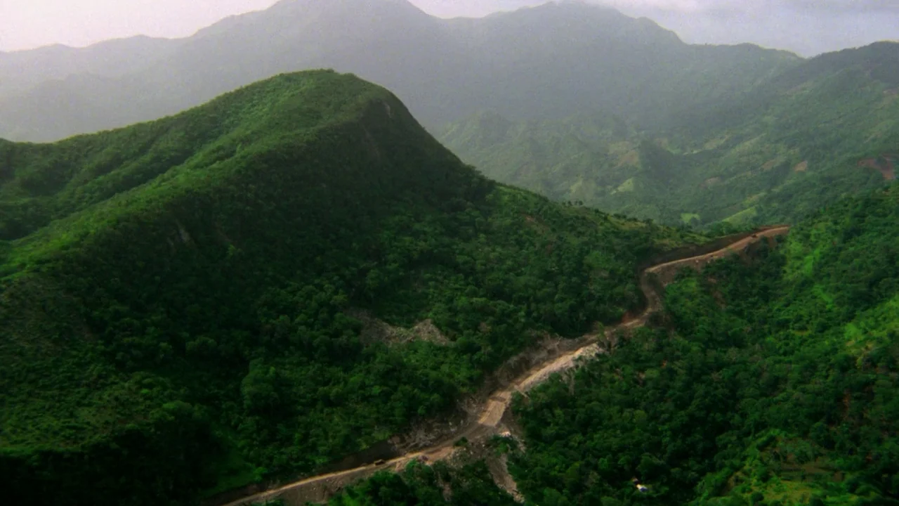 Sorcerer (1977) movie still: establishing — Lush green mountain slopes dominate the scene under a soft, overcast daylight sky. A winding dirt track…