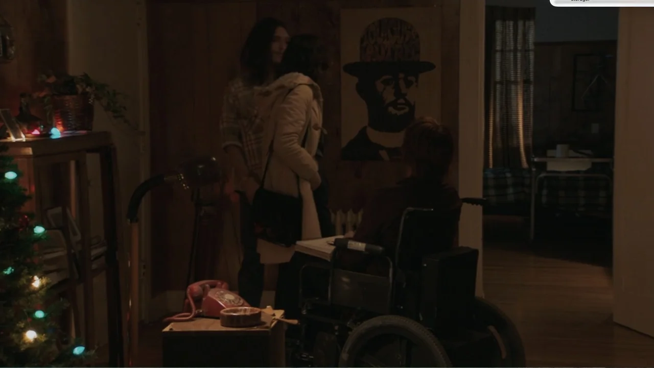 Don't Worry, He Won't Get Far on Foot (2018) movie still: medium — A dimly lit room with wood-paneled walls features a person in a wheelchair facing…