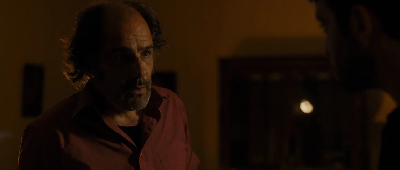 Road Games (2015) movie still: medium — A dark, dimly lit room with barely visible details in the background. A middle-aged man stands in the…