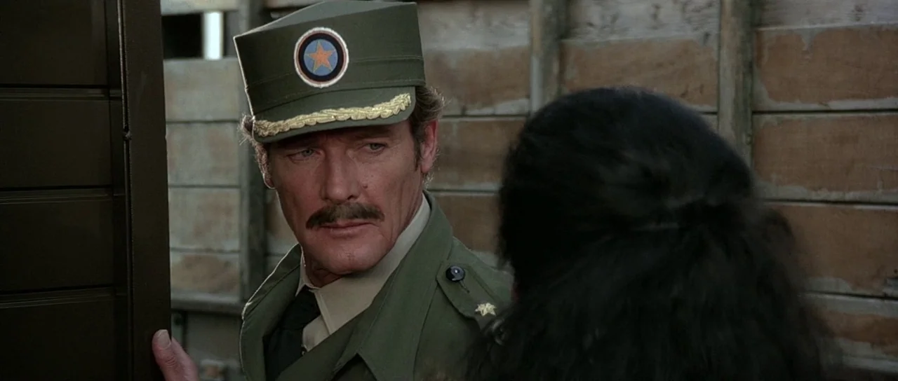 Octopussy (1983) movie still: closeup — A man in a military uniform stands inside the wooden bed of a truck. He looks at a woman whose back is…