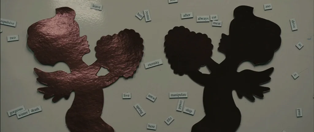 Pontypool (2008) movie still: closeup — A white magnetic board features two dark cherub silhouettes and various scattered word tiles. The words…