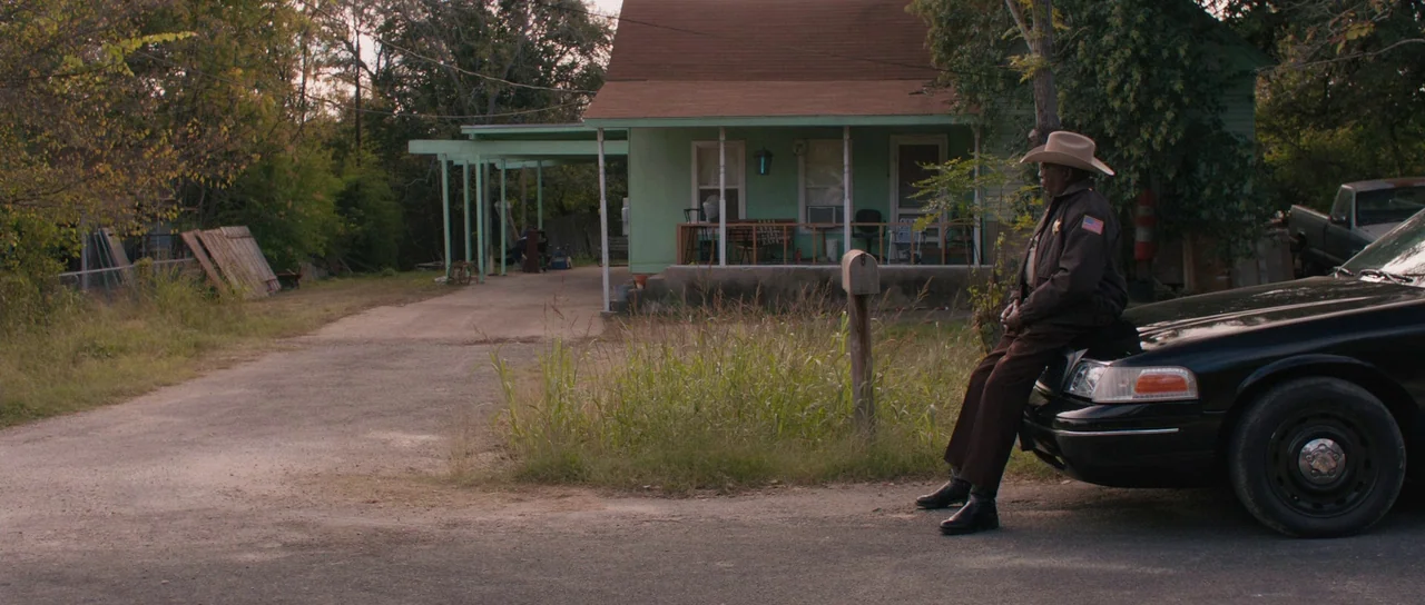 Joe (2013) movie still: wide — A rural setting features a small green house with a front porch and gravel driveway. An elderly sheriff sits…