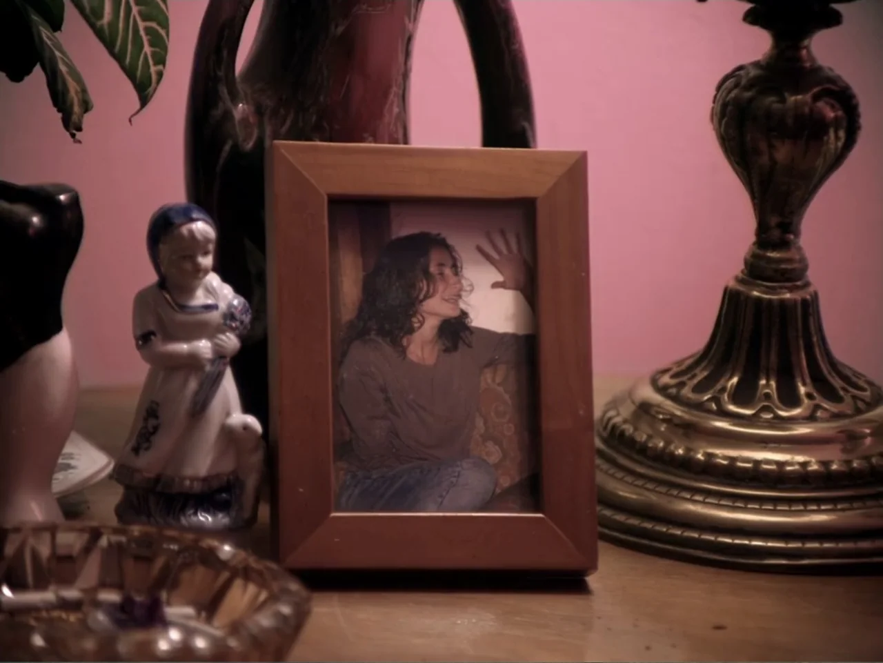 History and Geography (2024) movie still: medium — A wooden picture frame sits on a table amongst various household knick-knacks. Inside the photo,…