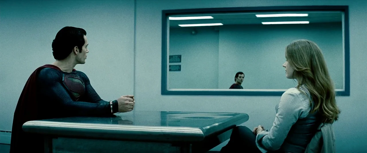 Man Of Steel (2013) movie still: medium — An austere room with a metal table and a large observation window on the wall. A man in a blue costume and…