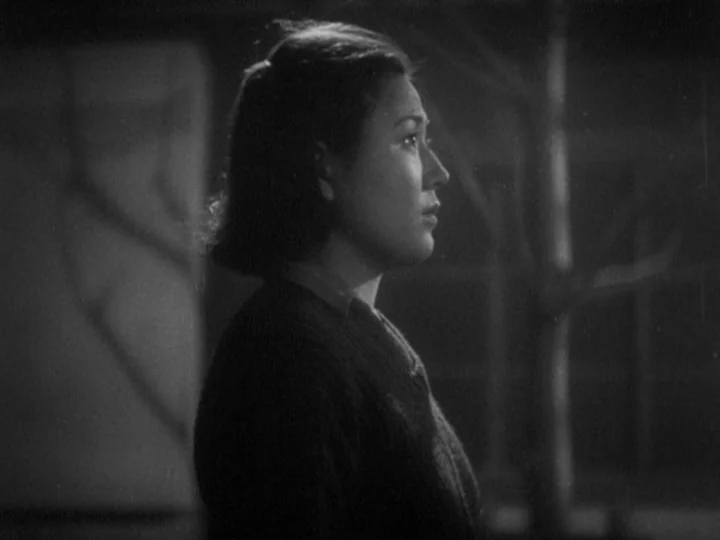 The Most Beautiful (1944) movie still: medium — A dark outdoor space at night with a blurred tree silhouette visible behind the woman. The woman…