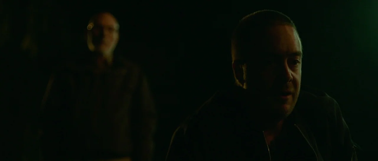 Green Room (2015) movie still: medium — Two men stand in a dark, outdoor environment at night. The foreground man is sharp while the man in the…
