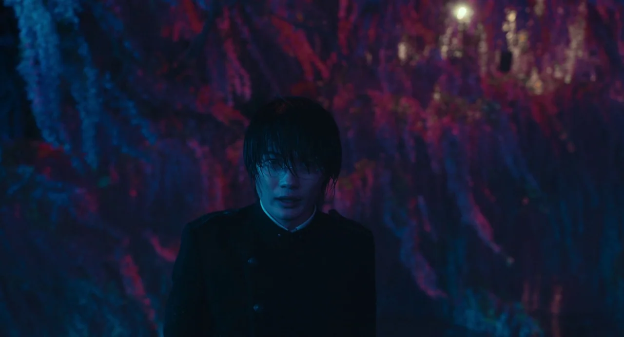 xxxHolic (2022) movie still: medium — A dark environment populated by dense clusters of illuminated hanging wisteria flowers. A young man stands…