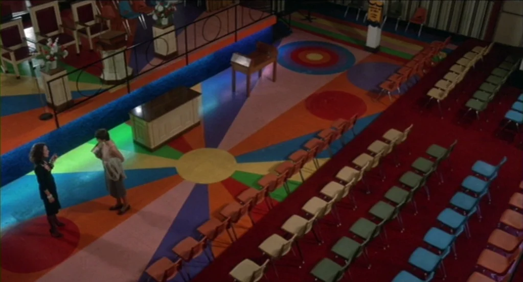Cat People (1982) movie still: wide — A community center features a brightly painted floor with large circular patterns and rows of chairs. Two…