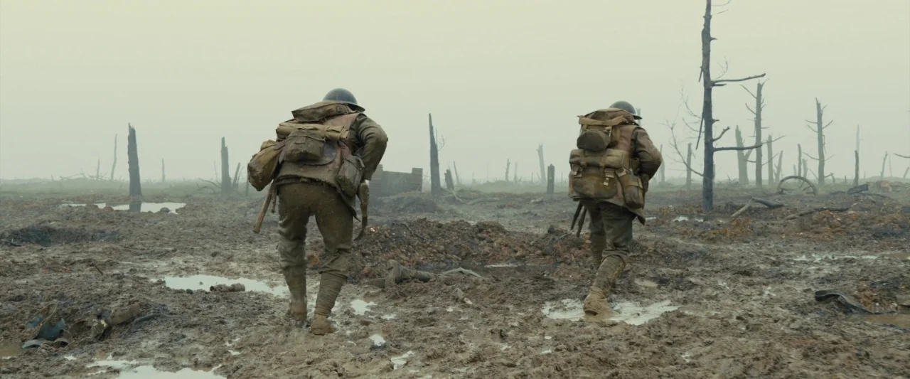 1917 movie still: fullbody — A desolate, mud-covered wasteland is filled with puddles and leafless charred tree trunks. Two soldiers in uniforms and…