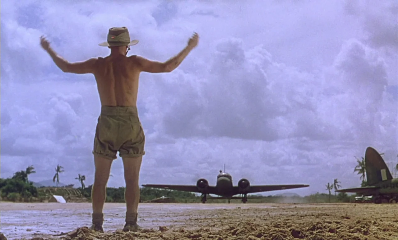 The Purple Plain (1954) movie still: fullbody — An open dirt landing strip in a tropical area with palm trees and sky. A man stands facing away from…