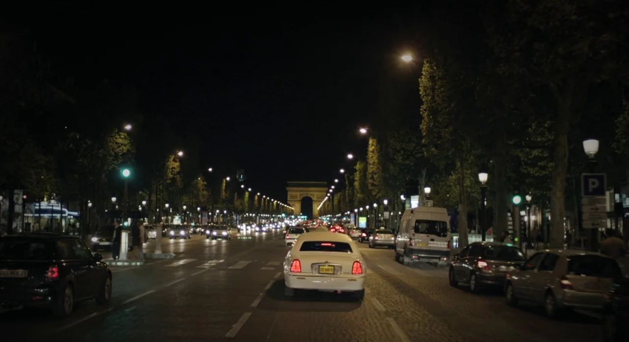 Holy Motors (2012) movie still: wide — A wide city avenue in Paris is lined with mature trees at night. The Arc de Triomphe is visible in the…