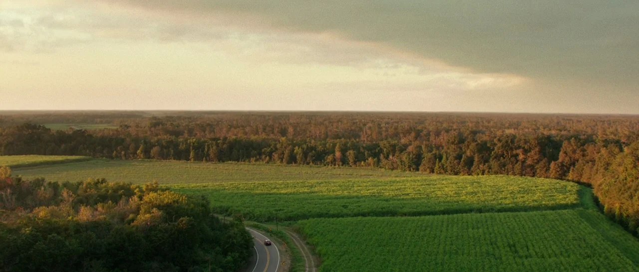 10 Cloverfield Lane (2016) movie still: establishing — A vast expanse of lush green crop fields stretches toward a distant dense forest. A winding…