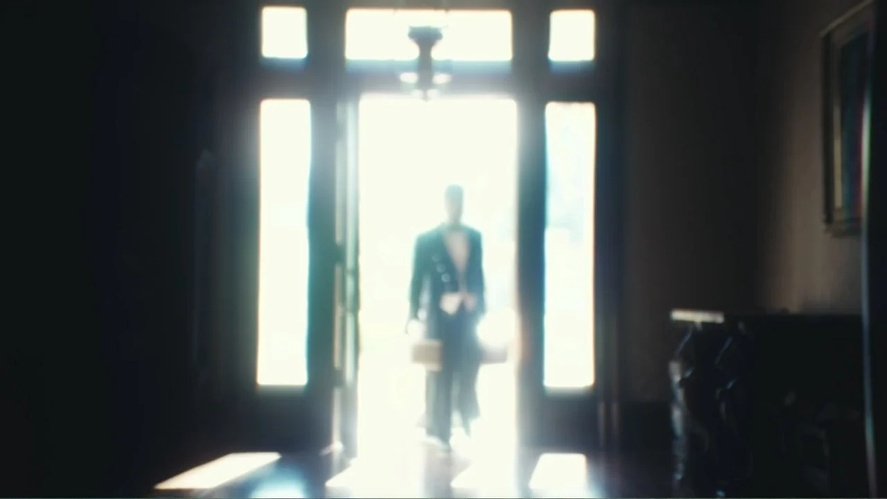 Demon in the White House (2021) movie still: fullbody — A dark, blurred hallway leads toward a bright open doorway into a light space. A man in a…